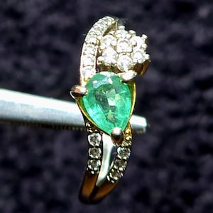Puede incluir: Un anillo de oro con una esmeralda en forma de pera y un grupo de diamantes más pequeños. El anillo tiene una banda delicada y retorcida.