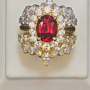 May include: A gold ring featuring a large, oval-cut, red gemstone surrounded by smaller, clear gemstones. The ring has a halo design with additional clear gemstones and a gold band. The ring is displayed on a white surface.