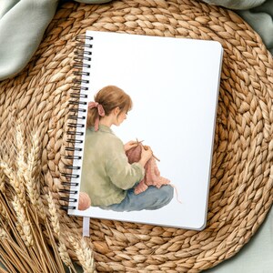 May include: A spiral-bound notebook featuring a watercolour illustration of a person knitting. The person wears a green shirt and blue trousers, with a pink bow in their hair. A ball of yarn sits nearby. The notebook rests on a woven mat.