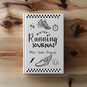 Może przedstawiać: Biały dziennik biegacza z czarnym tekstem i ilustracjami. Okładka zawiera słowa "Haley's Running Journal" oraz frazy "Miles • Goals • Progress". Ilustracje obejmują but do biegania, stoper i uśmiechniętą buzię.