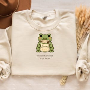 May include: Cream-colored sweatshirt featuring a cartoon frog holding a jar labeled "Starter." The text "emotionally attached to my starter" is below the frog. The sweatshirt has a crew neck and long sleeves.