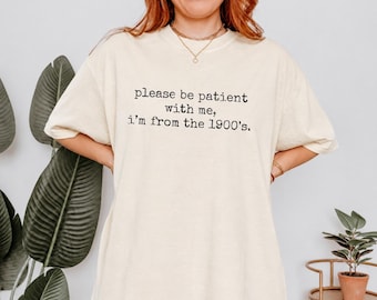May include: An off-white short-sleeve t-shirt featuring black typewriter-style text on the chest that reads, please be patient with me, i'm from the 1900's. The shirt has a crew neckline and a relaxed fit, made from a solid light-colored fabric.