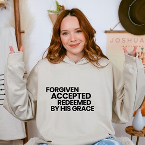 Pode incluir: Um moletom bege com as palavras "FORGIVEN ACCEPTED REDEEMED BY HIS GRACE" em letras pretas. O moletom tem capuz com cordão e mangas compridas. A pessoa que usa o moletom está sorrindo e fazendo o sinal de paz.