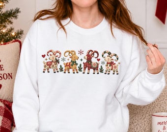 May include: A white crewneck sweatshirt featuring a graphic print of four stylised goats decorated with festive patterns, red and green scarves, and bows, interspersed with small red hearts and snowflakes across the chest area.