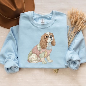 May include: A pale blue sweatshirt with a Cavalier King Charles Spaniel dog illustration. The dog has a patchwork design with floral and gingham patterns. The dog has brown and white fur and is sitting.