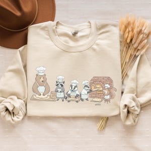 May include: A cream-coloured sweatshirt with a charming illustration of animals baking bread. A bear, raccoon, badger, and squirrel are shown wearing chef hats and aprons, with a brick oven in the background. The design is playful and suitable for casual wear.