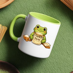 May include: White ceramic mug with a green handle and interior. The mug features a cartoon frog holding a jar labeled "Starter" with a wooden spoon and bowl. The design is on a green background.