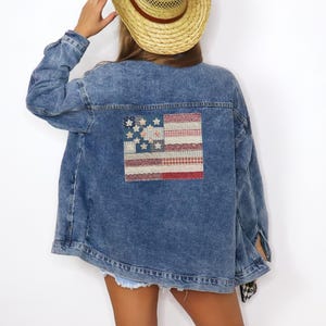 May include: A blue denim jacket with a patchwork American flag design on the back. The flag features stars and stripes in red, white, and blue fabrics. The jacket has a classic collar and button-down front.