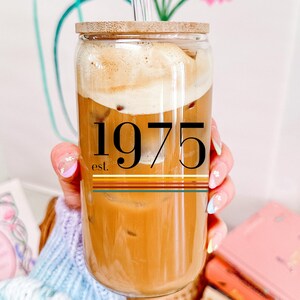 May include: A glass iced coffee beverage in a clear glass with a bamboo lid and a straw. The glass has a black and white graphic that says "est. 1975" with colorful stripes.