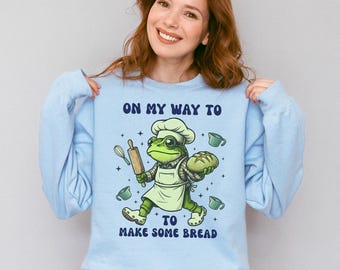 Op de afbeelding: Lichtblauwe sweatshirt met een cartoon kikkerkok met brood en een deegroller. De kikker draagt een koksmuts en een schort, met de tekst "ON MY WAY TO MAKE SOME BREAD". Het ontwerp bevat bakgerelateerde iconen zoals maatbekers en een garde.