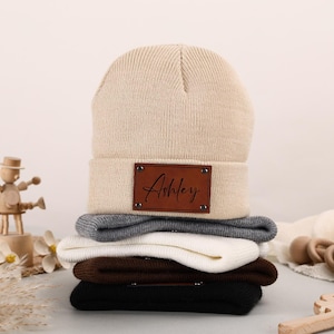 Puede incluir: Una pila de gorros de punto en varios colores, incluyendo beige, gris, blanco, marrón y negro. El gorro superior tiene un parche de cuero marrón con el nombre "Ashley" en cursiva. Los gorros están hechos de material de punto.
