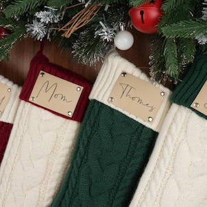Puede incluir: Cuatro calcetines navideños de punto trenzado en rojo, crema y verde, cada uno con una etiqueta de cuero personalizada. Las etiquetas dicen "Rachel", "Mom", "Thomas" y "Dad". Decoración festiva navideña con ramas de hoja perenne y adornos.