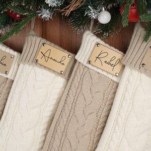 May include: Four cable-knit Christmas stockings in cream and beige hang from a wooden surface, each with a personalized leather tag. The tags are inscribed with names: Sophia, Amanda, Rachel, and Henry. A paw print is next to Henry's name.