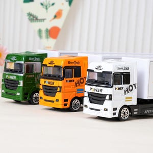 May include: Three toy semi-trucks in green, orange, and white. Each truck has the word "POWER" on the front and "HOT" on the side. The white truck has a white trailer with a lightning bolt and the word "KRU".
