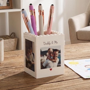 Puede incluir: Un portalápices blanco con fotos y el texto "Daddy & Me" contiene varios bolígrafos de varios colores. El portalápices está sobre una mesa de madera, con una taza y un dibujo cerca. Los bolígrafos tienen detalles en oro rosa.