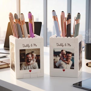 Peut inclure: Deux porte-stylos blancs avec une photo et le texte "Daddy & Me". Les supports sont remplis de stylos de différentes couleurs, dont or rose, gris et violet. Un cœur rouge est en dessous de la photo.