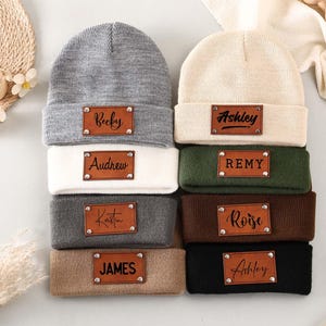 Puede incluir: Una pila de gorros personalizados en varios colores, incluyendo gris, blanco y negro. Cada gorro presenta un parche de cuero marrón con un nombre en escritura cursiva. Un accesorio de moda.