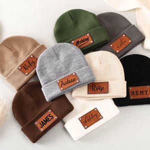 Può includere: Una collezione di berretti personalizzati in vari colori, tra cui beige, verde oliva, grigio e nero. Ogni berretto presenta una toppa in pelle marrone con un nome in un carattere scritto a mano. I berretti sono disposti su una superficie chiara.