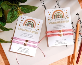 May include: Two matching bracelets on cards with the text "first day of KINDERGARTEN". The bracelets are pink and have a gold heart charm. The cards have a rainbow design and the text "Here are two bracelets, One Big and One Small For your First Day of School."