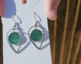 May include: A pair of dangle earrings with a silver-tone metal teardrop frame. Each earring features a circular, shimmering green center. The earrings are displayed on a white earring card.