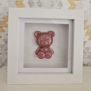 Puede incluir: Escultura de oso de peluche rosa brillante enmarcada. El oso está centrado en un marco de caja de sombra blanca. El fondo es blanco y el marco es blanco. El oso es de color sólido con una textura brillante.