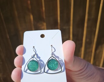 May include: A pair of dangle earrings with a teal glitter design. Each earring features a teal glitter circle set within a silver-colored triangle frame. The earrings are displayed on a white earring card.