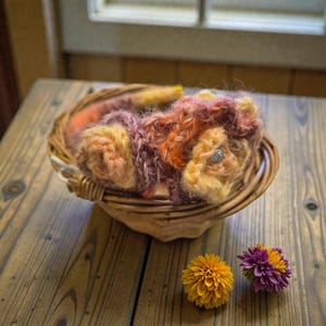 May include: A handmade, colorful headband with a floral design, resting in a woven basket. The headband features shades of orange, yellow, and purple yarn, with a sparkling embellishment on one of the flowers. Two decorative flowers sit beside the basket.