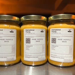 Può includere: Tre barattoli di vetro con coperchi dorati pieni di una salsa gialla. Ogni barattolo ha un'etichetta bianca con il testo "International Priority" e "House Chipotle Sauce".