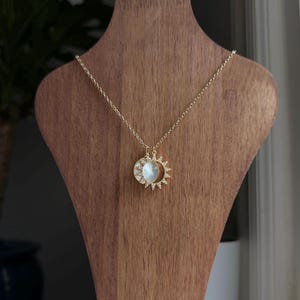 May include: A gold-colored necklace featuring a pendant with a sun and moon design. The pendant has a white, iridescent center with a sunburst and crescent moon outline. The necklace is displayed on a wooden jewelry stand.