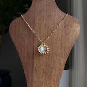 Puede incluir: Un collar de tono dorado con un colgante circular que presenta una incrustación central de nácar iridiscente. El colgante está rodeado por un diseño de rayos de sol con pequeñas piedras claras engastadas, suspendido de una delicada cadena dorada.