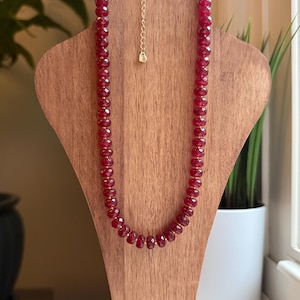 Può includere: Una collana di rubini sfaccettati esposta su un espositore di gioielli in legno. La collana presenta una serie di pietre preziose rosso intenso infilate con una catena color oro e un piccolo pendente. L'espositore è di colore marrone chiaro.