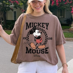 May include: Espresso-colored t-shirt featuring a vintage Mickey Mouse graphic. The shirt has a crew neck and short sleeves. The image also shows white shorts and a beige baseball cap. The overall style is casual and comfortable.