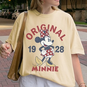 May include: Butter-colored t-shirt featuring a graphic of Minnie Mouse with the text "ORIGINAL 1928 MINNIE". The shirt is paired with white shorts and a white baseball cap. A brown shoulder bag is also visible.