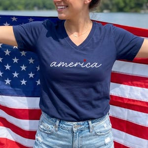 K&ouml;nnte beinhalten: Marineblaues V-Ausschnitt-T-Shirt mit dem Wort "america" in wei&szlig;er Schrift und einem roten Stern. Die Person h&auml;lt eine amerikanische Flagge. Das Shirt wird mit Jeansshorts kombiniert. Der Hintergrund umfasst einen See und B&auml;ume.