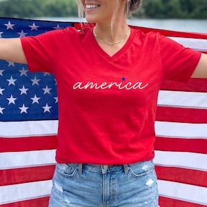 May include: A red V-neck t-shirt with the word "america" in cursive and a blue star. The shirt is paired with denim shorts. The background features an American flag and a scenic outdoor setting.