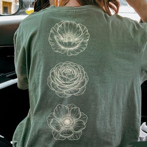 May include: A moss green t-shirt with three white floral illustrations on the back. The shirt is worn by a person in a car, and the text "SUPER SOFT" is visible on a pink sticker. The word "moss" is also visible.