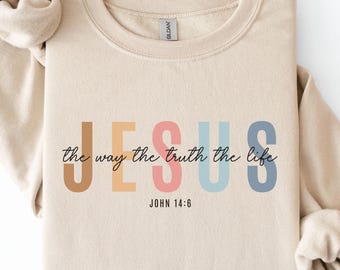 May include: A beige crewneck sweatshirt with the word "JESUS" in colorful block letters. Below the word "JESUS" is the phrase "the way the truth the life" in a script font. The text "JOHN 14:6" is printed below the script font.