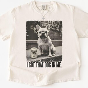 K&ouml;nnte beinhalten: Cremefarbenes T-Shirt mit einem Schwarz-Wei&szlig;-Bild einer franz&ouml;sischen Bulldogge mit Zigarette im Mund und einer Dose "Urban Paw Lager". Der Text "I GOT THAT DOG IN ME." ist unter dem Bild gedruckt.