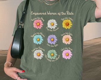 May include: An olive green t-shirt featuring the text "Empowered Women of the Bible" above a grid of nine colorful daisy illustrations, each paired with a name and a short phrase such as "Believe like Mary" and "Pray like Hannah."