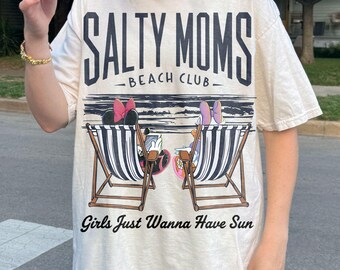 May include: Cream-colored t-shirt with the text "SALTY MOMS BEACH CLUB" in black. The shirt features a beach scene with two beach chairs and the phrase "Girls Just Wanna Have Sun" in black.