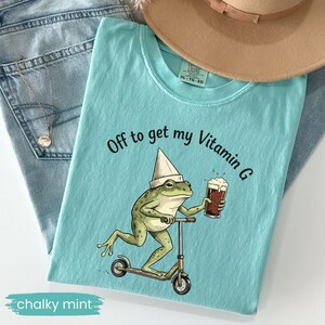 May include: A light blue t-shirt with the text "Off to get my Vitamin G" and a cartoon frog riding a scooter while holding a beer. The shirt is labeled "chalky mint".