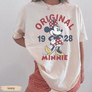 May include: Ivory-colored t-shirt featuring a vintage Minnie Mouse graphic. The design includes the words "ORIGINAL 1928 MINNIE" in red and blue, with Minnie Mouse wearing a red polka-dot bow and dress. The shirt is paired with red and white checkered shorts.