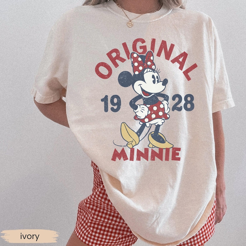 May include: Ivory-colored t-shirt featuring a vintage Minnie Mouse graphic. The design includes the words "ORIGINAL 1928 MINNIE" in red and blue, with Minnie Mouse wearing a red polka-dot bow and dress. The shirt is paired with red and white checkered shorts.