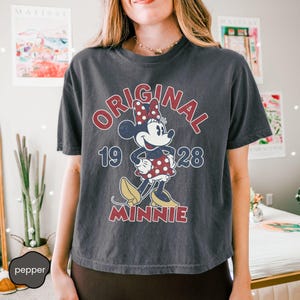 May include: Dark gray t-shirt featuring a vintage-style Minnie Mouse graphic. The design includes the words "ORIGINAL MINNIE 1928" in red and white, with Minnie Mouse wearing a polka-dot dress and bow.