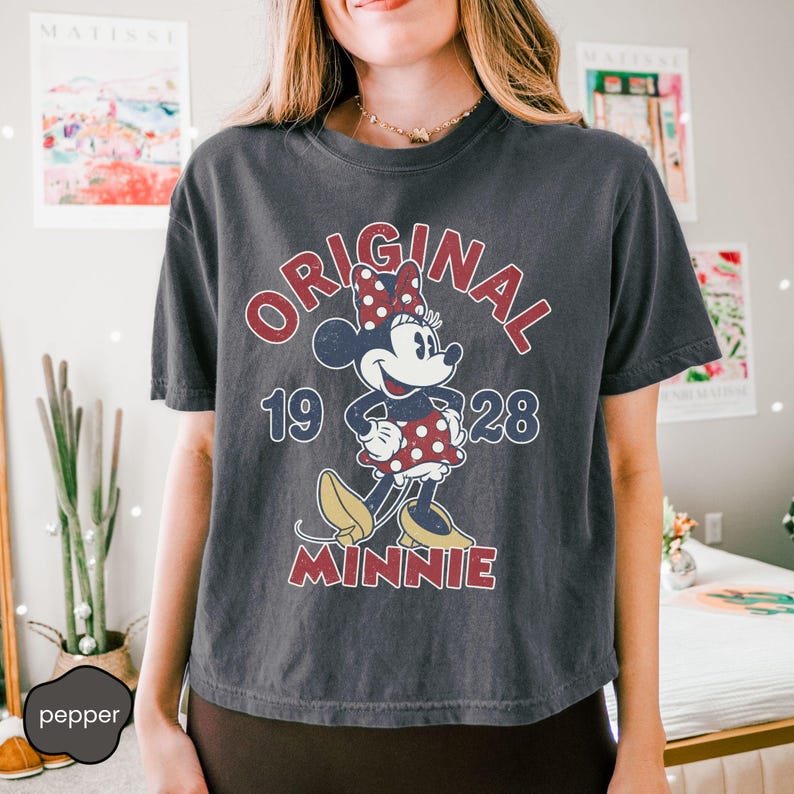 May include: Dark gray t-shirt featuring a vintage-style Minnie Mouse graphic. The design includes the words "ORIGINAL MINNIE 1928" in red and white, with Minnie Mouse wearing a polka-dot dress and bow.