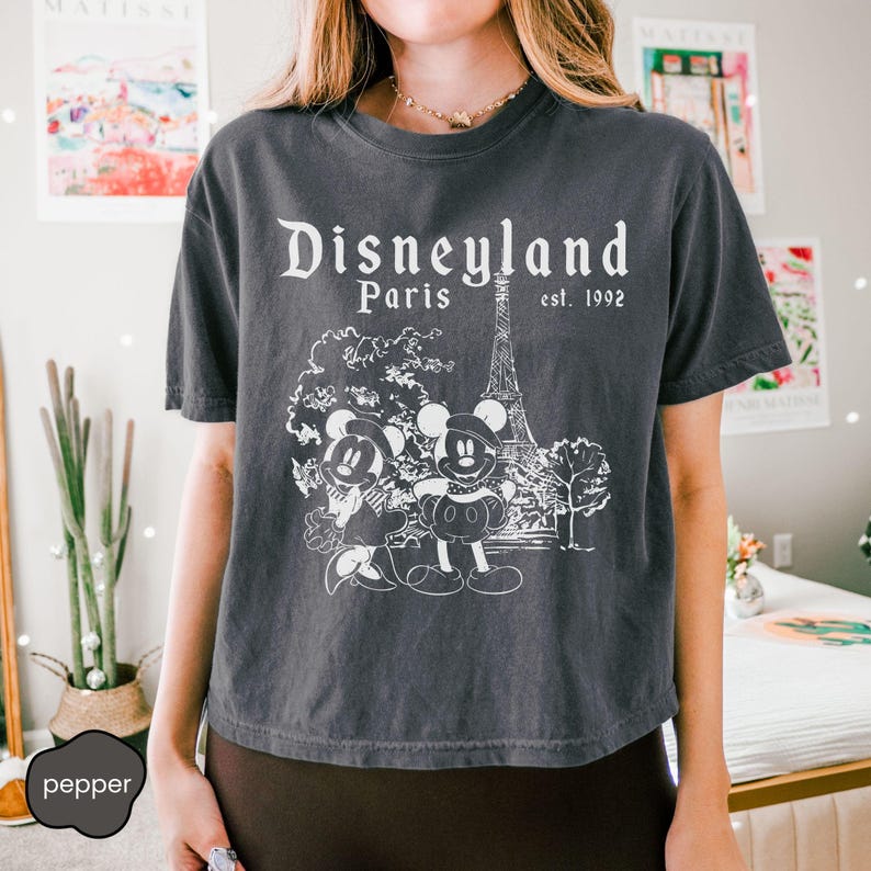 Puede incluir: Camiseta gris oscuro con el texto "Disneyland Paris est. 1992" y un dibujo de Mickey y Minnie Mouse frente a la Torre Eiffel. La camiseta tiene un aspecto vintage y desgastado.