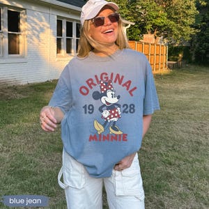 May include: A blue t-shirt featuring a Minnie Mouse graphic with the text "ORIGINAL 1928 MINNIE". The shirt has short sleeves and a crew neck. The graphic includes Minnie Mouse in a red and white polka dot dress.