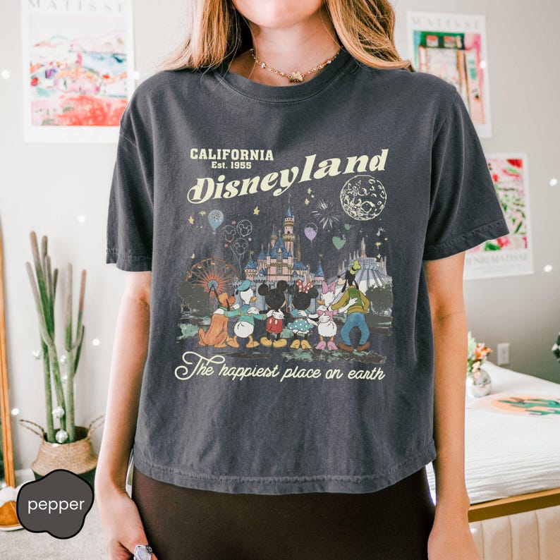 May include: Dark gray t-shirt featuring a Disneyland graphic. The design includes the text "California Est. 1955 Disneyland" with an illustration of the castle, characters, and fireworks. The phrase "The happiest place on earth" is also included.
