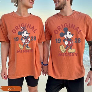 May include: Two orange t-shirts with the text "ORIGINAL" and the year "1928". One shirt features Minnie Mouse, the other Mickey Mouse. Both shirts are worn by people holding hands on a beach. The word "MINNIE" and "MICKEY" are below the characters.