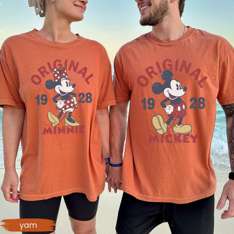 May include: Two orange t-shirts with the text "ORIGINAL" and the year "1928". One shirt features Minnie Mouse, the other Mickey Mouse. Both shirts are worn by people holding hands on a beach. The word "MINNIE" and "MICKEY" are below the characters.